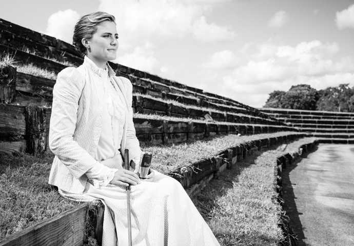 汤普森站在水边的草地上，手持球杆眺望，微风轻拂她的帽纱与衣裙，画面宁静而富有诗意，仿佛一位伫立在水一方的古典贵族，思考着下一杆的挥击。