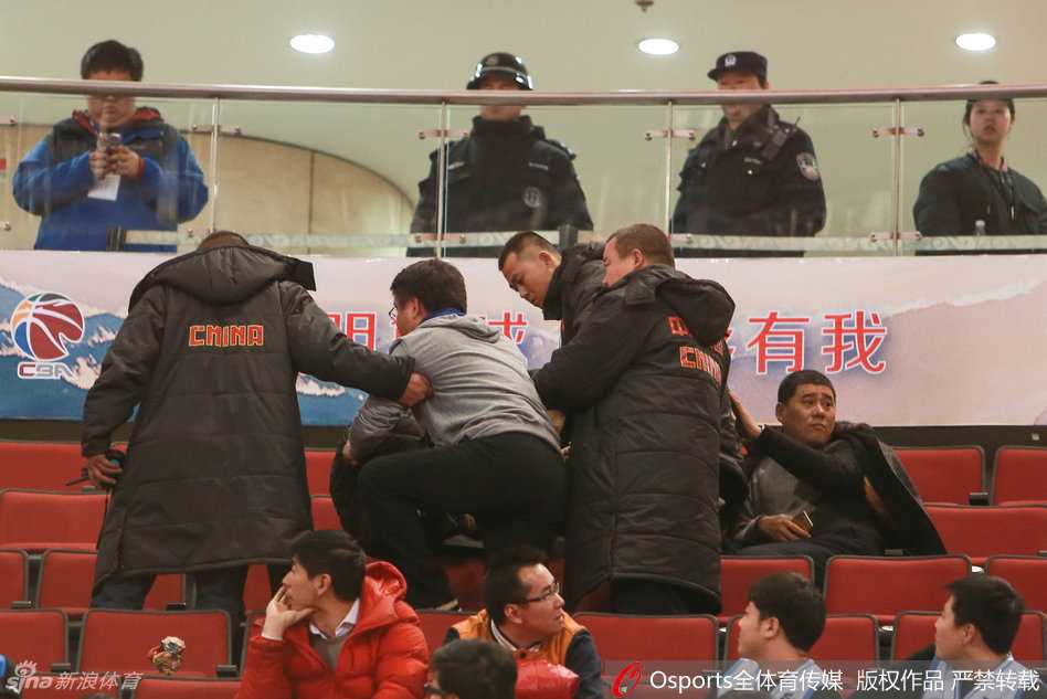 冲突现场，一名年轻球迷被同伴紧紧抱住，他试图挣脱束缚，脸上的表情交织着沮丧与愤怒。身后的看台上，更多的球迷站起身来，伸长脖子观望事态发展，现场气氛紧张到了极点。