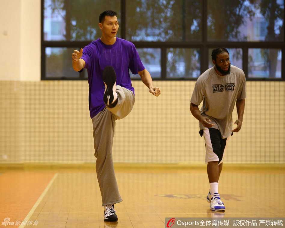 易建联手扶篮架进行踢腿拉伸，身体舒展到极致，一旁的陈江华侧身回望，脸上带着逗趣的表情正在说笑。