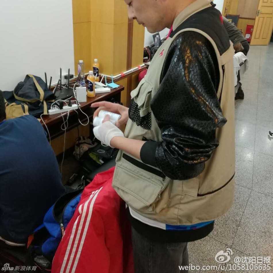 媒体工作室里，一位年纪较长的记者正低头看着手机，腕间的绷带在衣袖旁若隐若现。周围年轻的同行们同样手缠绷带，各自忙碌，整个空间弥漫着一种静谧而坚定的职业氛围。