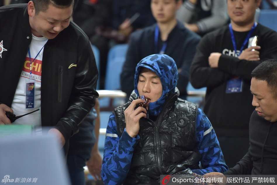 郭艾伦站起身在场边活动，厚实的外套显得他有些笨拙可爱，他依然不忘拿着那根棒棒糖，仿佛这是他在这个寒冷秋日里最重要的伙伴，脸上写满了“心情不错”。