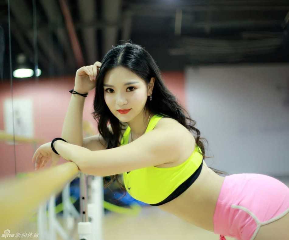 在健身房暖色调的灯光下，啦啦队员轻轻侧身回眸，乌黑的长发随之飘动，展现出万种风情。她弓形身材充满力量感，健康的小麦色皮肤在光影下闪着微光，仿佛凝固了时光。