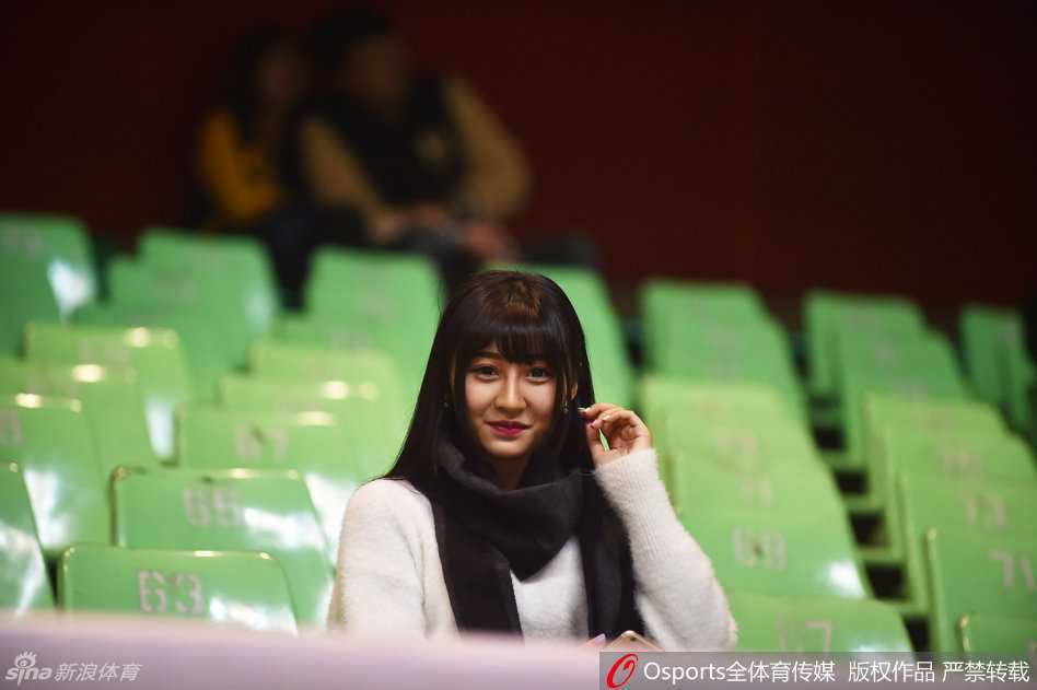 一位身着黑色时尚外套的温婉女球迷，面对镜头露出羞怯而欣喜的笑容，仿佛在为自己的主队暗暗祈祷