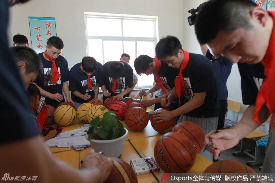 辽篮岫岩公益行 李晓旭与孩子们共筑篮球梦 李晓旭弯腰俯身,面带微笑地为一名系着红领巾的小学生在篮球上签名,周围聚满了充满期待眼神的孩子们。