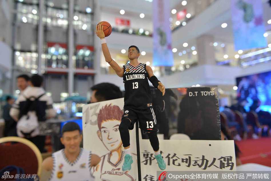 郭艾伦身穿黑色T恤端坐在签售台前，正低头在定制人偶的包装盒上认真签名，周围聚满了举着手机拍照的粉丝，桌上整齐摆放着待售的人偶礼盒。