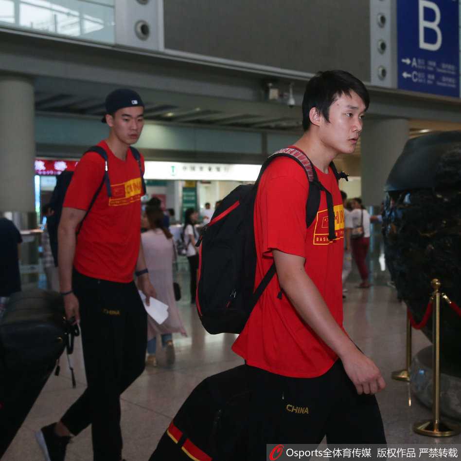 老将周鹏神情轻松地推着行李，身边的李慕豪显得憨厚朴实，背景是首都机场熙攘的人群。