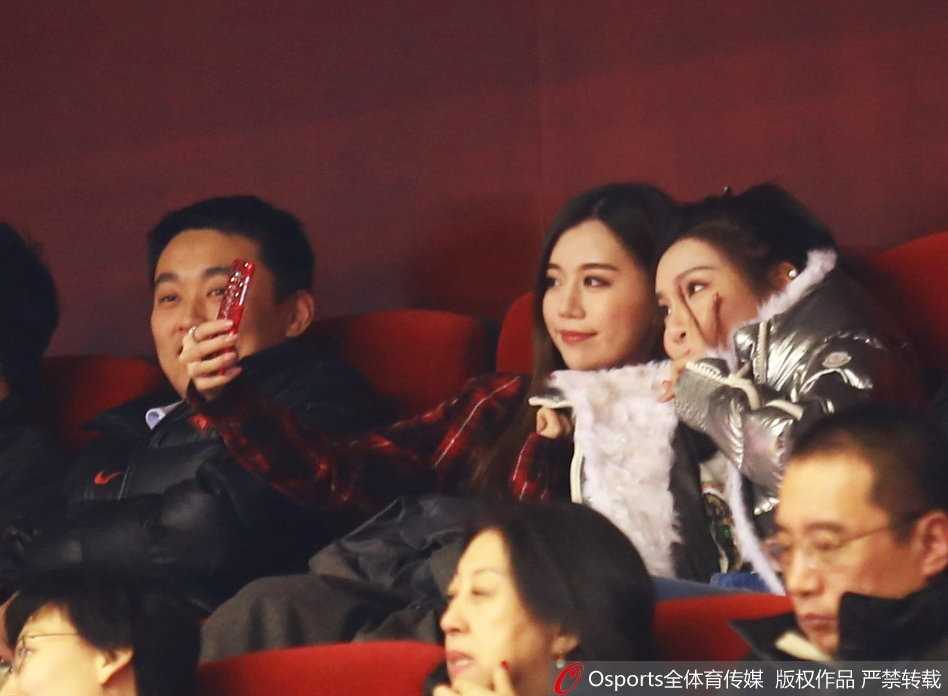 身披山东队应援外套的丁彦雨航女友与睢冉妻子在看台上并肩而坐，两人对着镜头露出灿烂笑容，仿佛春日暖阳