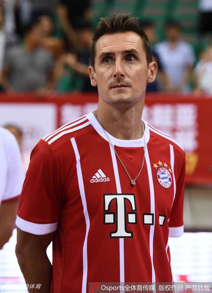 镜头拉近，克洛泽与现场主持人谈笑风生，他深邃的眼眸中带着好奇与亲切，倾听关于这场篮球赛的介绍，背景是色彩斑斓的斯坦科维奇杯赛场。