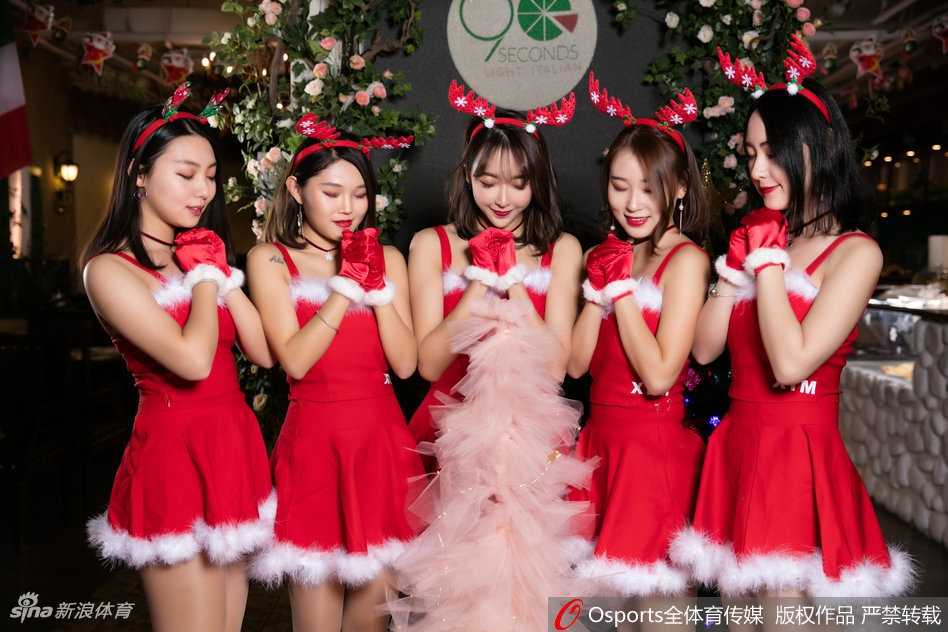 两位身着圣诞装的啦啦队员在摆满礼物的桌子后亲密合影，她们不仅准备了实用的帽子等小礼品，更用青春洋溢的笑容感染着过往的每一位球迷，现场充满了欢声笑语。