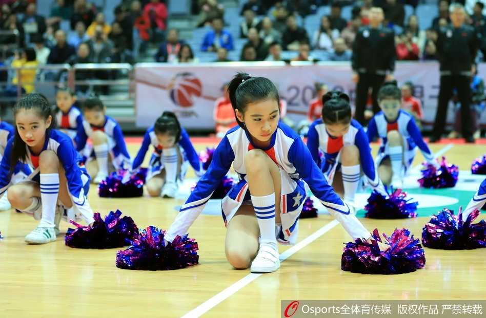 几位扎着丸子头的小宝贝身穿红色球衣，手拿金色花球，踮起脚尖认真观看比赛，眼神里满是好奇与兴奋