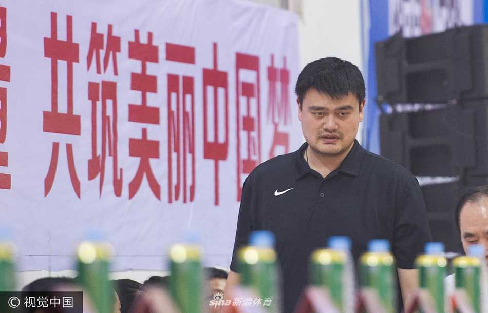 在喧闹的体育馆内，姚明侧身倾听身旁友人的话语，表情认真而专注，偶尔点头回应，眼神中透露出对场上局势的关切与思考。