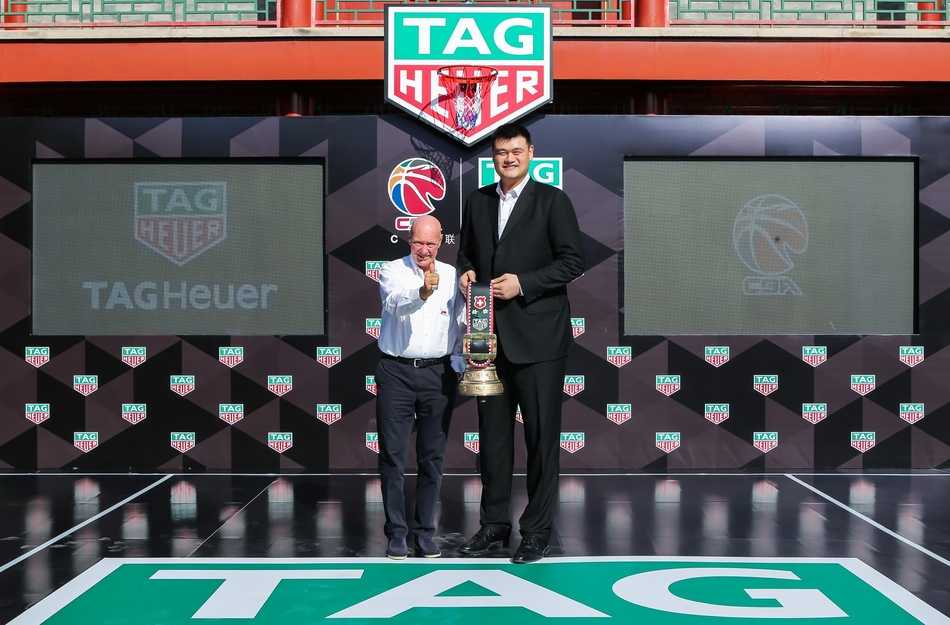 在北大朗润园万众楼前，姚明身着深色西装，面带微笑，目光坚定地注视前方，其身后是古色古香的建筑飞檐，象征着CBA传统与创新的交融。