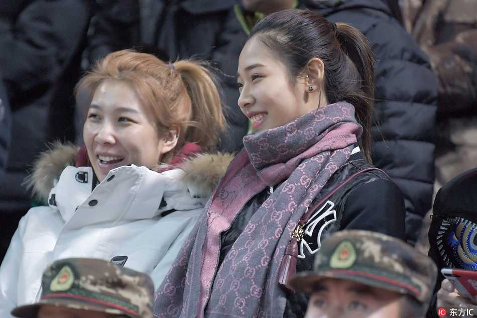 两位长发披肩的美女球迷并肩而坐，其中一位穿着时尚的迷彩外套，正与身旁的伙伴低声耳语，脸上洋溢着观看比赛特有的兴奋笑容。