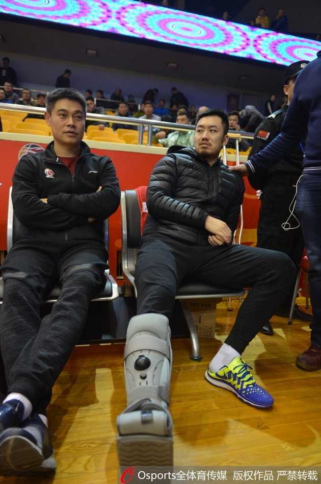 睢冉低着头坐在板凳席末端，左脚的保护靴被抬高搭在一个箱子上，他手中拿着一瓶水，表情显得有些落寞，与身后庆祝的球迷形成了鲜明对比。