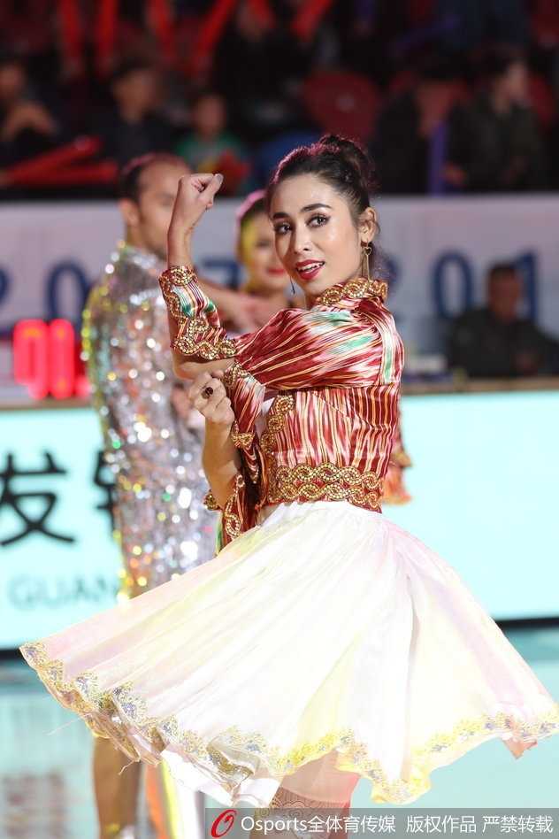 这位新疆美女穿着具有民族特色的服饰，头戴精致的小花帽，面带羞涩又大方的微笑，向观众挥手致意，极具亲和力。