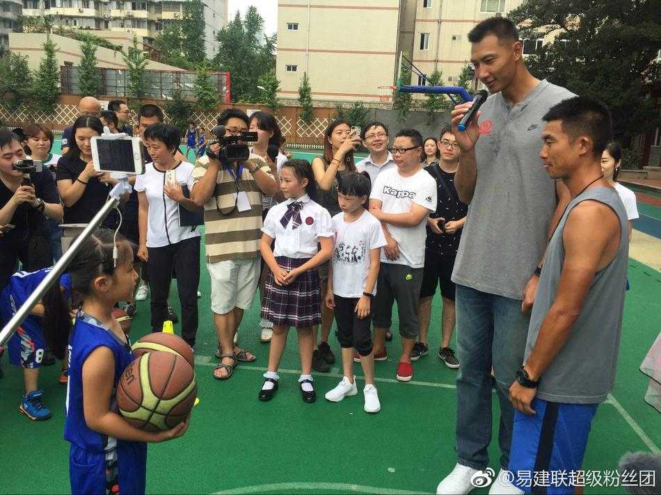 几位小面试官坐在课桌后神情专注，其中一位戴着红领巾的男孩正拿着话筒向站在对面的易建联提问。易建联双手背在身后，微微俯身侧耳倾听，仿佛真的在认真接受一场工作面试。