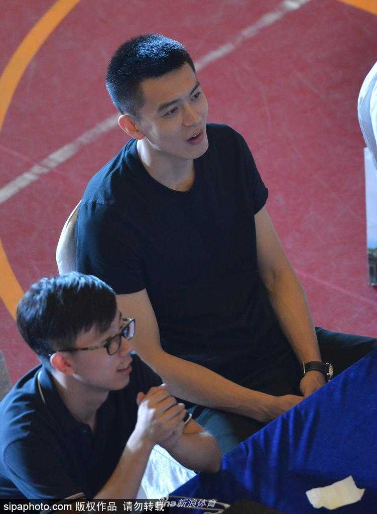 近距离特写下，杨鸣的笑容极具感染力，眉眼弯弯，眼神清澈而坚定。他手持话筒，似乎在分享着什么有趣的篮球故事，身后球迷的目光齐刷刷地聚焦在他身上。