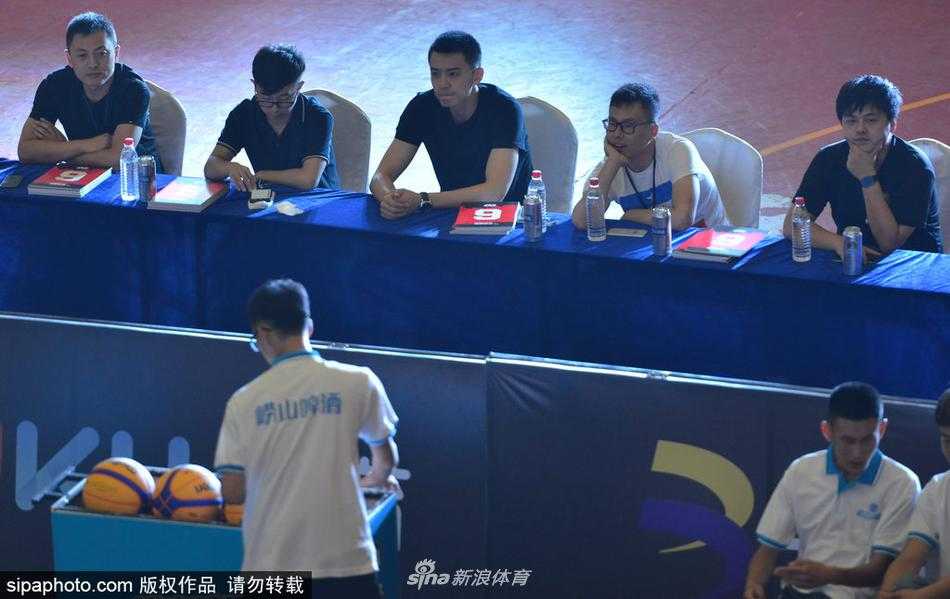 杨鸣坐在评委席上，手拿打分表，神情专注而认真。他身后的背景是热闹的三人篮球赛场，彩旗飘扬，而他仿佛隔绝了周围的喧嚣，沉浸在专业的评判之中。