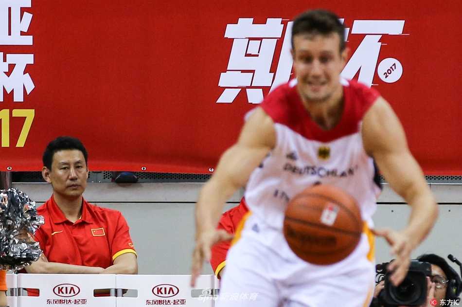 全景视角下，中国队的替补席沿着场边一字排开。红色的座椅、红色的球衣，构成一片红色的海洋。球员们的坐姿各异，但头部转向的方向惊人一致——都朝着球场中央。远处，德国与克罗地亚的球员正在拼抢，激烈的对抗与此刻场边的静态观摩形成鲜明对比，仿佛两个平行世界在这一刻通过篮球产生了交集。