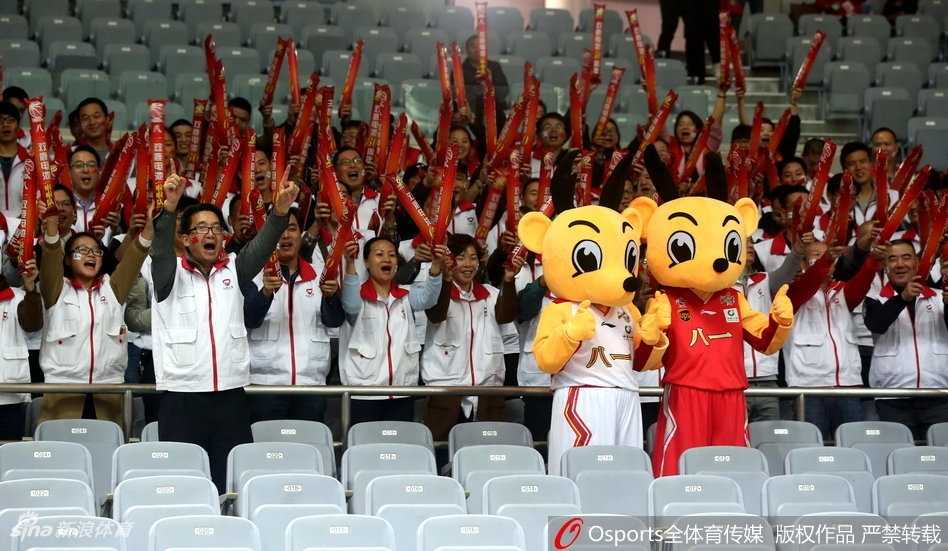 吉祥物站在看台台阶上向二楼的观众抛洒小礼物，它仰着头用力挥舞着手中的纪念品，灯光照亮了它身上红色的球衣，上方无数双手臂高高举起，仿佛一片欢呼的森林