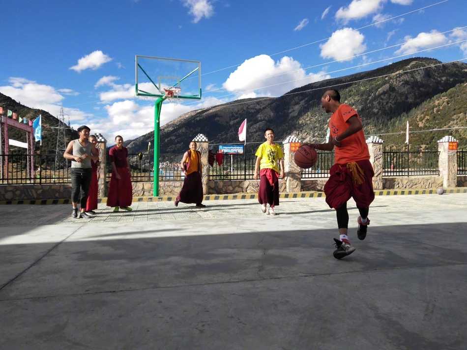 一名年轻的喇嘛正准备投篮，他双腿微曲，双手持球举过头顶，僧袍的衣摆垂落在地。在他身后，是连绵的雪山和湛蓝的天空，构成一幅动静结合、充满禅意的画面。