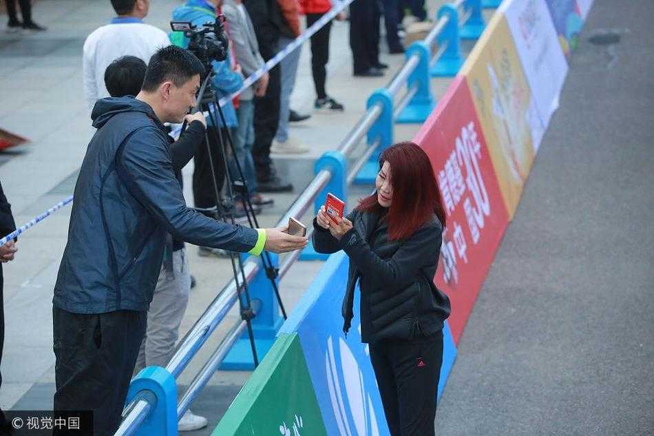 冬日娜身披央视外套，微笑着拿出手机，杜锋则弯下腰凑近屏幕，两人在喧闹的马拉松赛场边专注地扫描二维码，背景是模糊的人群和赛事横幅，画面充满了人情味。