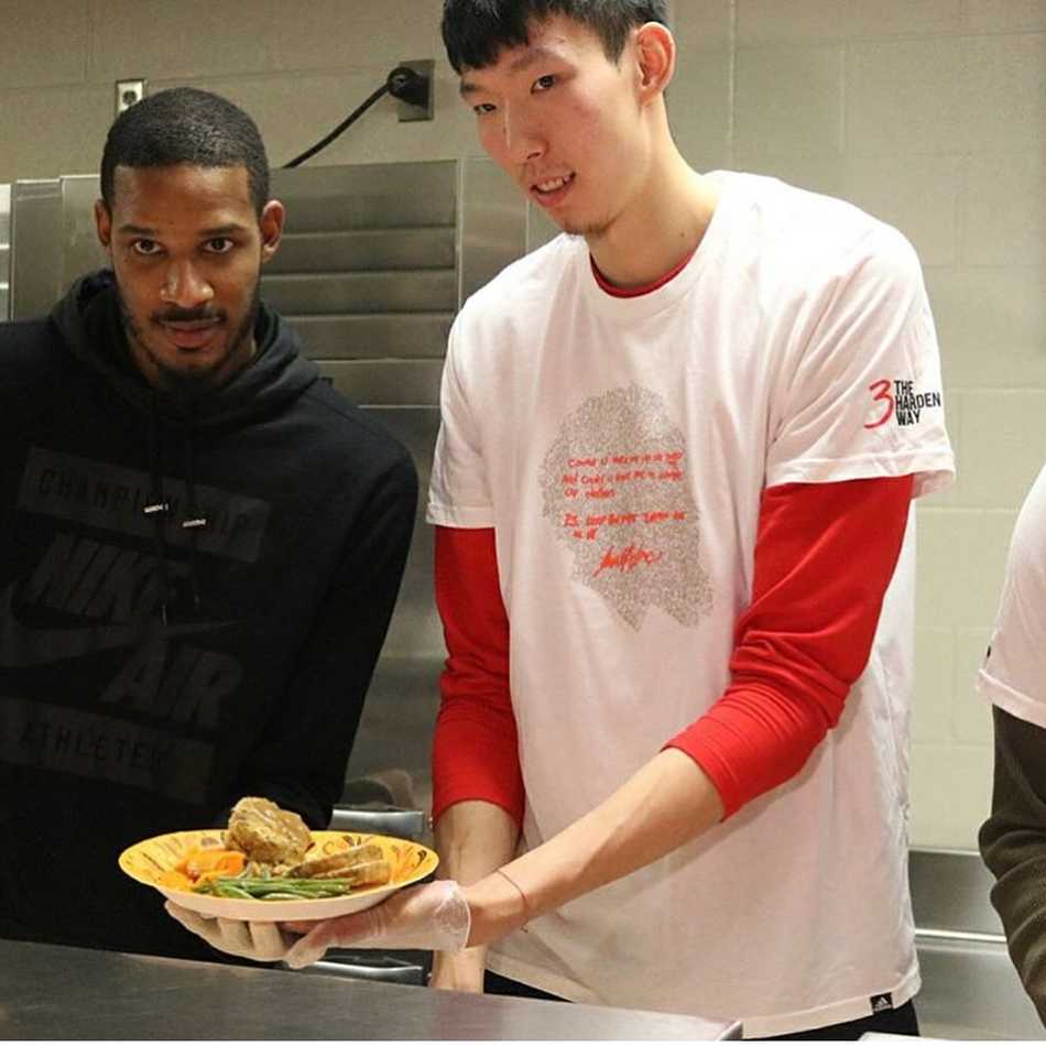 周琦与阿里扎手持餐盘，对着镜头做出搞怪的表情，两人身后是摆放整齐的节日餐点，整个画面充满了队友间的默契与欢乐。