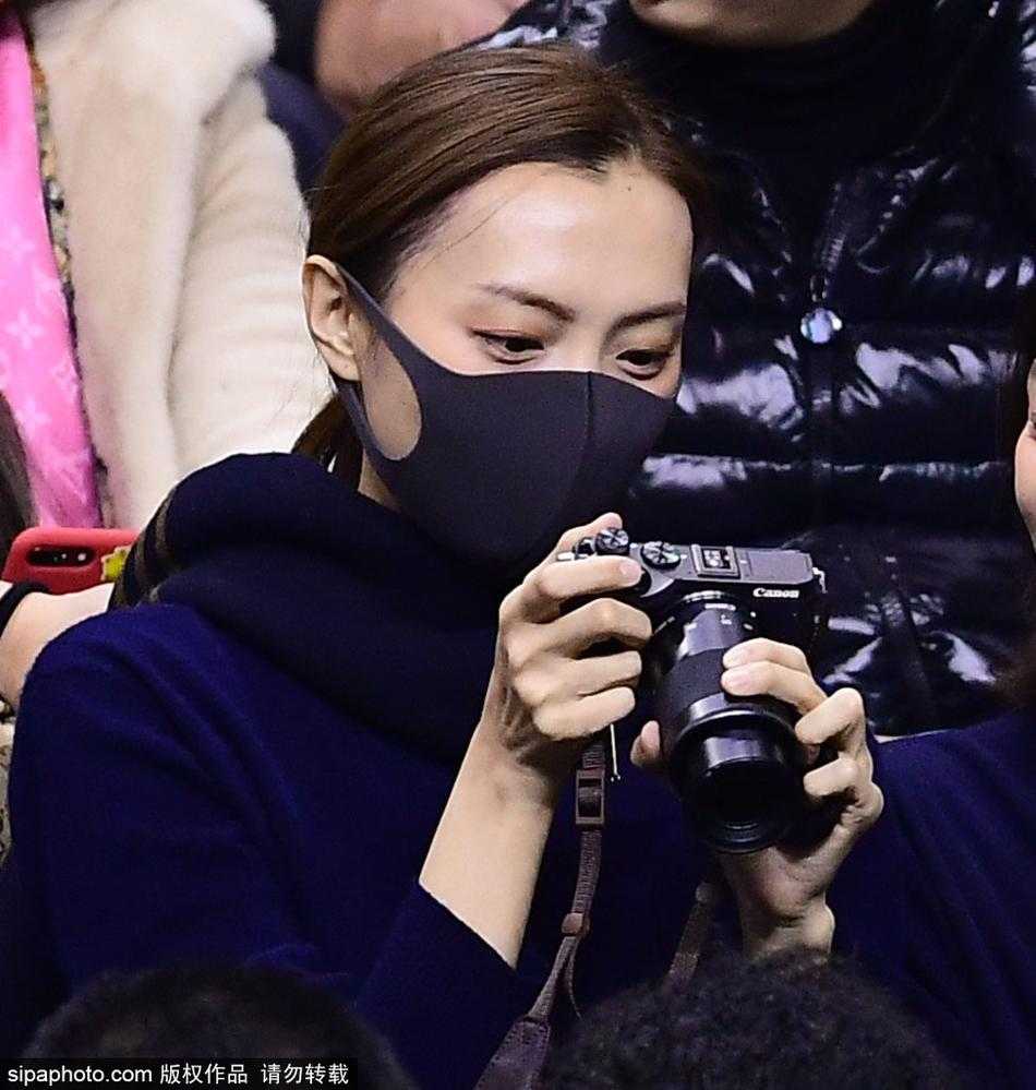 李旭身披大衣，手持专业相机，神情专注地站在场边，镜头紧紧追随着球场上的丈夫李晓旭