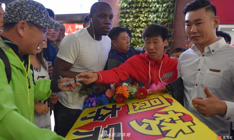 哈德森被热情的接机人群团团围住，他低头为几位幸运的球迷签名，闪光灯将他的脸庞照得格外清晰，仿佛定格了这荣耀的一刻。