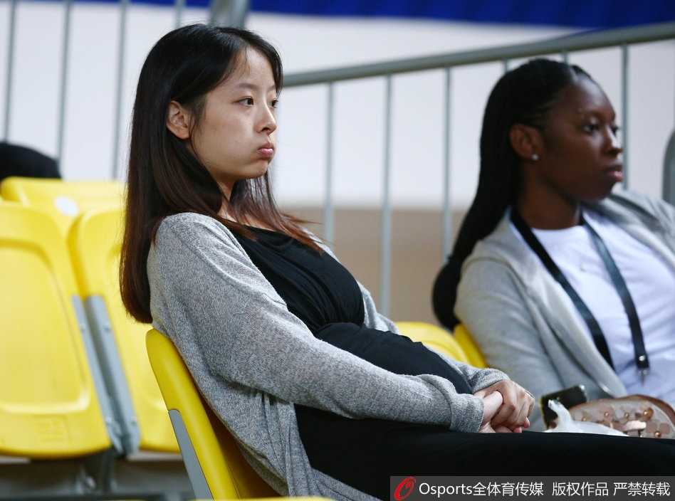 孙桐林妻子身着宽松衣物，双手轻抚在小腹前，眼神温柔地望向赛场方向，脸上洋溢着幸福而含蓄的微笑。