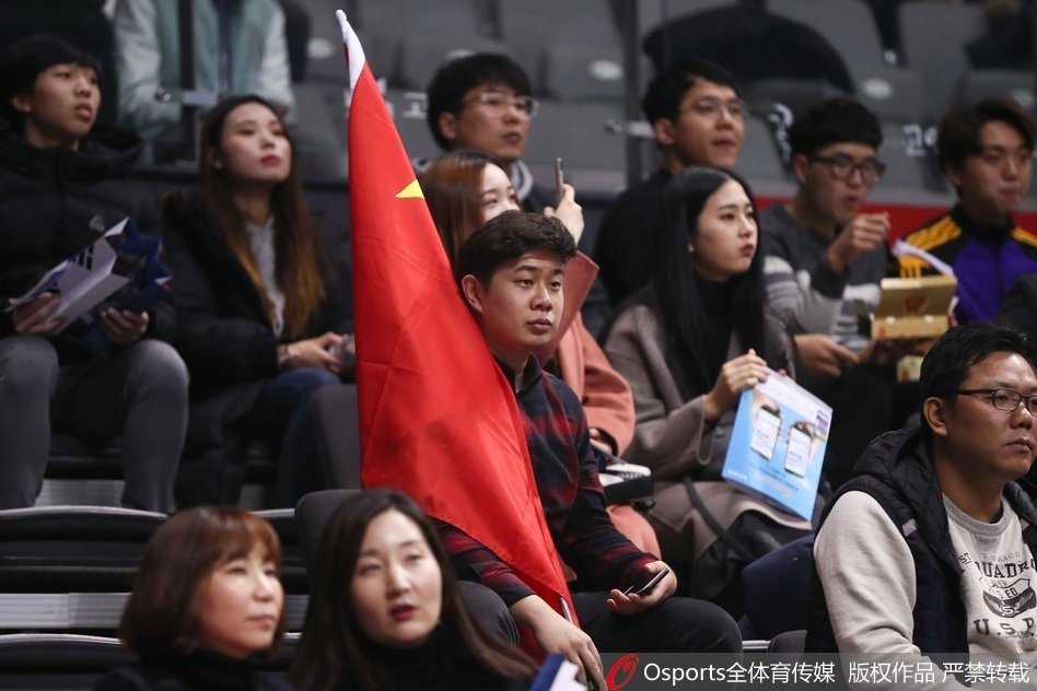 在韩国主场高阳体育馆的看台上，一名中国球迷神情专注且骄傲，双手奋力展开一面巨大的五星红旗，旗帜如火焰般在异国他乡的球馆内舒展。