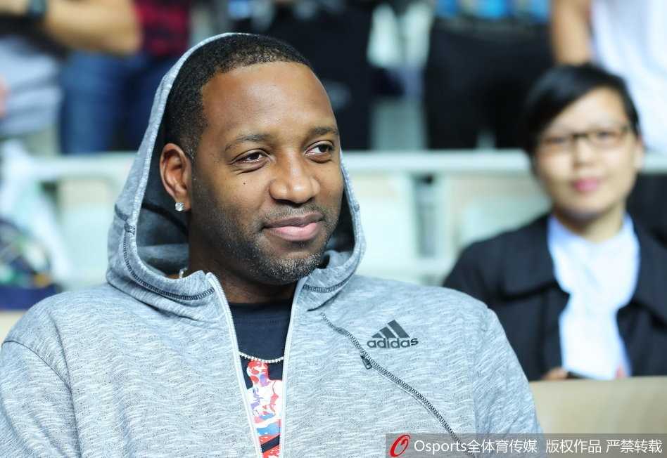 麦蒂现身源深体育馆,昔日得分王微笑见证CBA经典一战 麦蒂的侧脸特写,轮廓分明,他微微侧身看向球场另一侧,背景是虚化的观众席和灯光,营造出一种虽置身人群却独享回忆的宁静感。